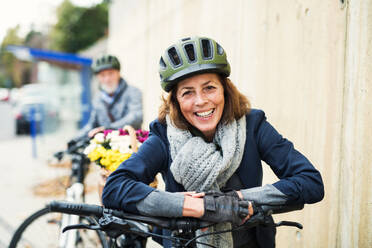 Active senior couple with helmets and electrobikes standing outdoors on a pathway in town. - HPIF28690