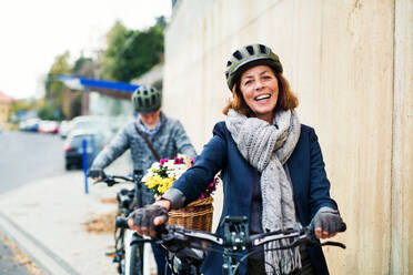 Active senior couple with helmets and electrobikes standing outdoors on a road in town. - HPIF28692