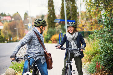 An active senior couple with helmets and electrobikes standing outdoors on a pathway in town, looking at each other. - HPIF28701