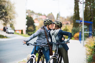 Active senior couple with helmets and electrobikes standing outdoors on a road in town, kissing. - HPIF28703