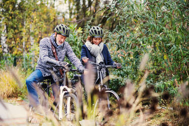 An active senior couple with helmets and electrobikes standing outdoors on a road in nature, looking at speedometer. - HPIF28704