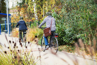 A rear view of active senior couple with helmets and electrobikes cycling outdoors on a pathway in town. - HPIF28709