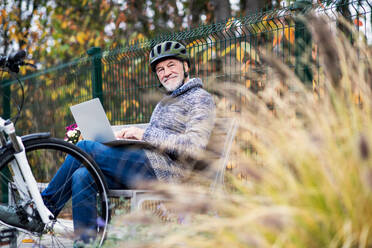 A senior man with electrobike sitting on a bench outdoors in town in autumn, using laptop. Copy space. - HPIF28711