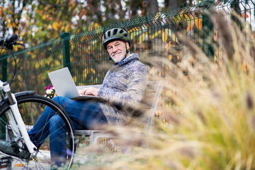 Ein älterer Mann mit Elektrofahrrad sitzt auf einer Bank im Freien in der Stadt im Herbst und benutzt einen Laptop. Platz kopieren. - HPIF28711