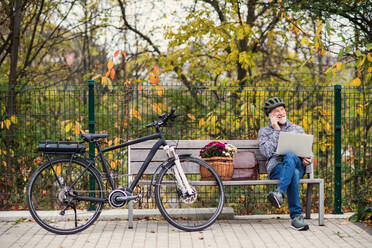 A senior man with electrobike, laptopand smartphone sitting on a bench outdoors in town in autumn, making a phone call. Copy space. - HPIF28712