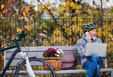 A senior man with electrobike, laptop and smartphone sitting on a bench outdoors in town in autum. Copy space. - HPIF28713