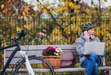 Ein älterer Mann mit Elektrofahrrad, Laptop und Smartphone sitzt im Herbst auf einer Bank in der Stadt. Kopierraum. - HPIF28713