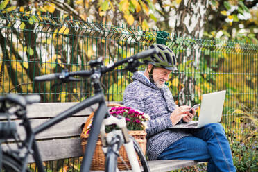 Ein älterer Mann mit Elektrofahrrad, Laptop und Smartphone sitzt im Herbst auf einer Bank in der Stadt. Kopierraum. - HPIF28714