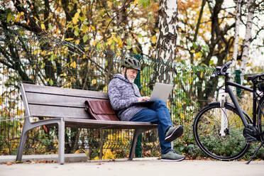 Ein älterer Mann mit Elektrofahrrad sitzt auf einer Bank im Freien in der Stadt im Herbst und benutzt einen Laptop. Platz kopieren. - HPIF28715