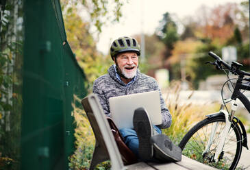 Ein älterer Mann mit Elektrofahrrad sitzt auf einer Bank im Freien in der Stadt im Herbst und benutzt einen Laptop. Platz kopieren. - HPIF28716