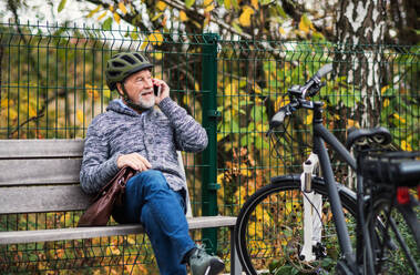 Ein älterer Mann mit Elektrofahrrad und Smartphone sitzt im Herbst auf einer Bank im Freien in der Stadt und führt ein Telefongespräch. - HPIF28718