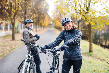 Ein aktives Seniorenpaar mit Helmen und Elektrofahrrädern, das draußen auf einer Straße in der Natur steht. - HPIF28730