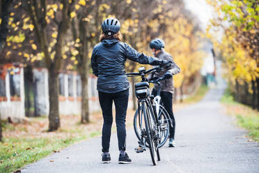 Rückansicht eines aktiven Seniorenpaares mit Helmen und Elektrofahrrädern, das im Freien auf einer Straße im Park steht. - HPIF28738