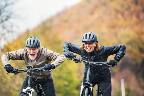 An active senior couple with helmets and electrobikes cycling outdoors on a road in nature. - HPIF28745