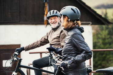An active senior couple with helmets and electrobikes standing outdoors in front of a house. - HPIF28747