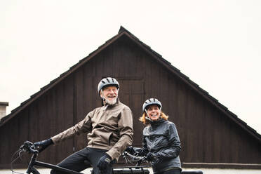 An active senior couple with helmets and electrobikes standing outdoors in front of a house. - HPIF28752