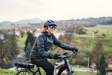 Seitenansicht einer aktiven älteren Frau mit Elektrofahrrad, die im Herbst im Freien radelt. - HPIF28768