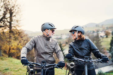 An active senior couple with helmets and electrobikes standing outdoors on a road in nature. - HPIF28769