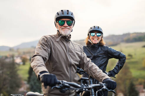 An active senior couple with helmets and electrobikes standing outdoors on a road in nature. - HPIF28770