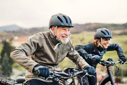 An active senior couple with helmets and electrobikes cycling outdoors on a road in nature. - HPIF28774