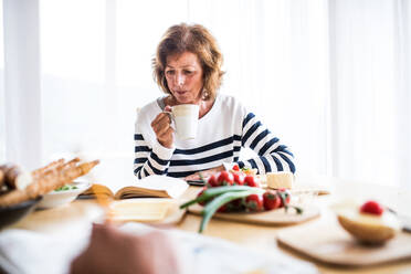 Ältere Frau frühstückt zu Hause. Eine alte Frau sitzt am Tisch und liest ein Buch. - HPIF28829
