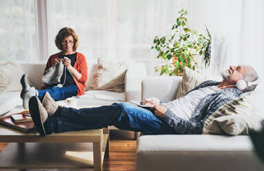 Happy senior couple relaxing at home. A woman knitting and a man with tablet listening to music. - HPIF28842