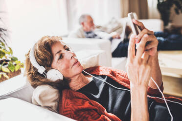 Happy senior couple relaxing at home. A woman with smartphone and headphones listening to music. - HPIF28845