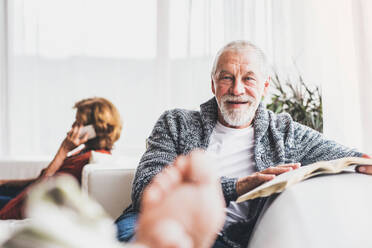 Happy senior couple with smartphone relaxing at home. A woman making a phone call and a man reading a book. - HPIF28849