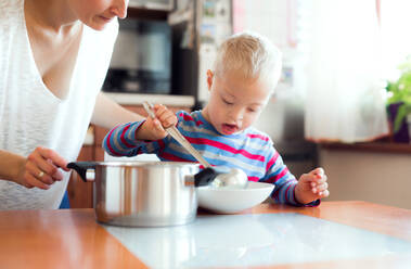 Ein glücklicher behinderter Junge mit Down-Syndrom und einer nicht erkennbaren Mutter, die ihm zu Hause einen Teller Suppe einschenkt, zur Mittagszeit. - HPIF28919