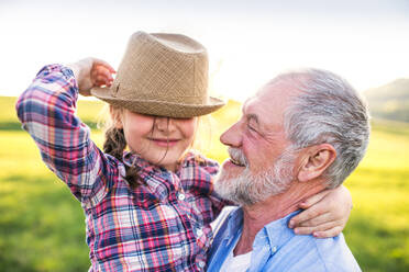 A cheerful small girl with grandfather outside in spring nature, having fun with a hat. - HPIF28983