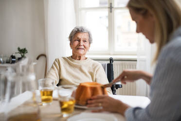 Eine ältere Frau im Rollstuhl mit einer Gesundheitsberaterin sitzt zu Hause am Tisch und schneidet einen Kuchen an. - HPIF29084