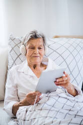 A sick senior woman with headphones and tablet lying in bed at home or in hospital, listening to music. - HPIF29137