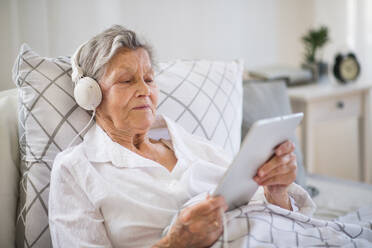 A sick senior woman with headphones and tablet lying in bed at home or in hospital, listening to music. - HPIF29138