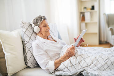 A sick senior woman with headphones and tablet lying in bed at home or in hospital, listening to music. - HPIF29142
