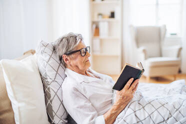 A sick senior woman with eyeglasses lying in bed at home or in hospital, reading bible book. - HPIF29144