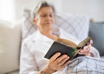 A sick senior woman lying in bed at home or in hospital, reading bible book. - HPIF29145