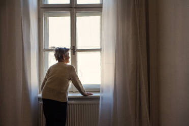 Rückansicht einer älteren Frau, die zu Hause steht und aus dem Fenster schaut; Kopierraum. - HPIF29153