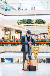 A senior man with bags standing in a shopping center at Christmas time, checking the time. - HPIF29171