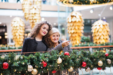 Ein Porträt einer älteren Großmutter und ihrer Enkelin im Teenageralter, die zur Weihnachtszeit in einem Einkaufszentrum stehen und auf etwas zeigen. - HPIF29215