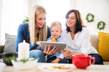Ein kleiner Junge mit Mutter und Großmutter sitzt zur Weihnachtszeit zu Hause auf einem Sofa und benutzt ein Tablet. - HPIF29235