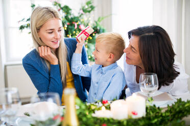 Ein kleiner Junge mit Geschenk und Mutter und Großmutter, die zur Weihnachtszeit zu Hause an einem Tisch sitzen. - HPIF29246