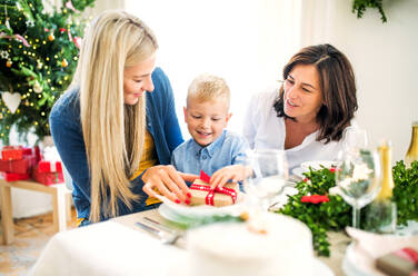 Ein kleiner Junge mit Geschenk und Mutter und Großmutter, die zur Weihnachtszeit zu Hause an einem Tisch sitzen. - HPIF29247