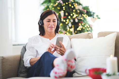 Eine ältere Frau mit Kopfhörern und Smartphone sitzt zu Hause auf dem Sofa und hört Musik zur Weihnachtszeit. - HPIF29251