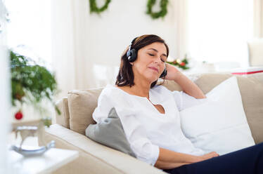 A senior woman with headphones and closed eyes sitting on a sofa at home, listening to music at Christmas time. - HPIF29254