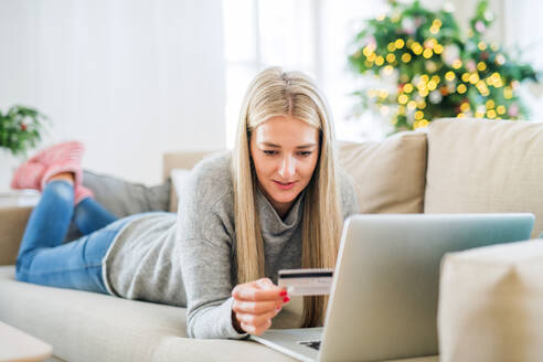 Eine junge Frau mit Kreditkarte und Laptop liegt zur Weihnachtszeit zu Hause auf dem Sofa und tätigt Online-Zahlungen. - HPIF29262