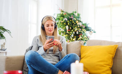 Eine junge Frau mit Kopfhörern und Smartphone sitzt zur Weihnachtszeit zu Hause auf dem Sofa und hört Musik. - HPIF29266