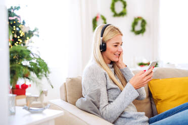 Eine junge Frau mit Kopfhörern und Smartphone sitzt zur Weihnachtszeit zu Hause auf dem Sofa und hört Musik. - HPIF29268