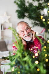 A cheerful senior man standing by Christmas tree, putting ball ornaments on his ears. - HPIF29298