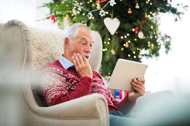 Ein überraschter älterer Mann sitzt auf einem Sessel am Weihnachtsbaum und benutzt ein Tablet. Festliche Zeit. - HPIF29309