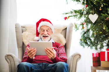 A portrait of senior man with Santa hat sitting on armchair at home at Christmas time, using tablet. - HPIF29311